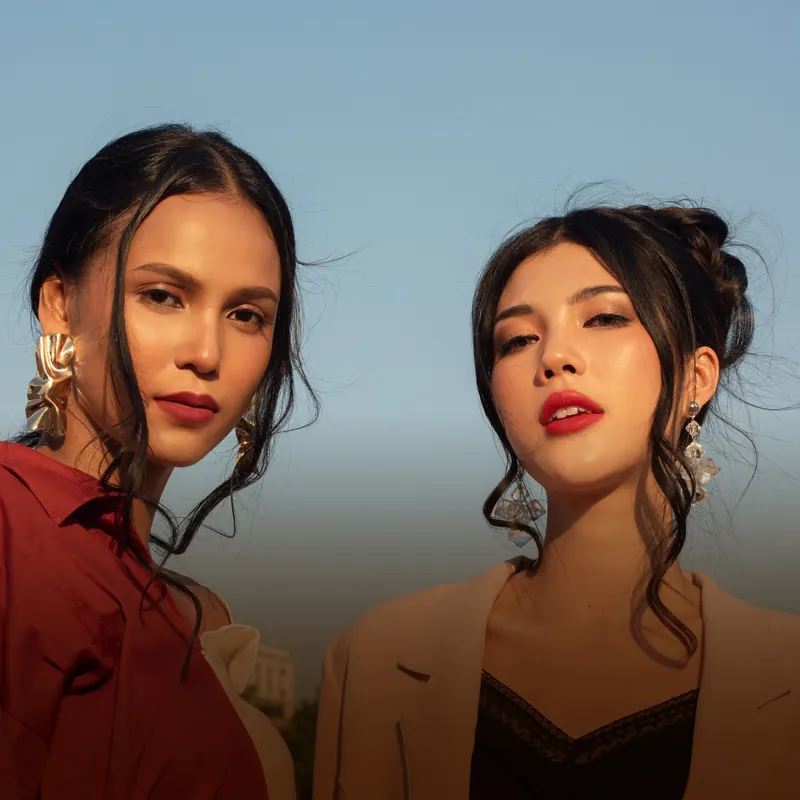 Dua model dengan riasan flawless natural, kemeja merah bata dan blazer krem, berlatar langit biru