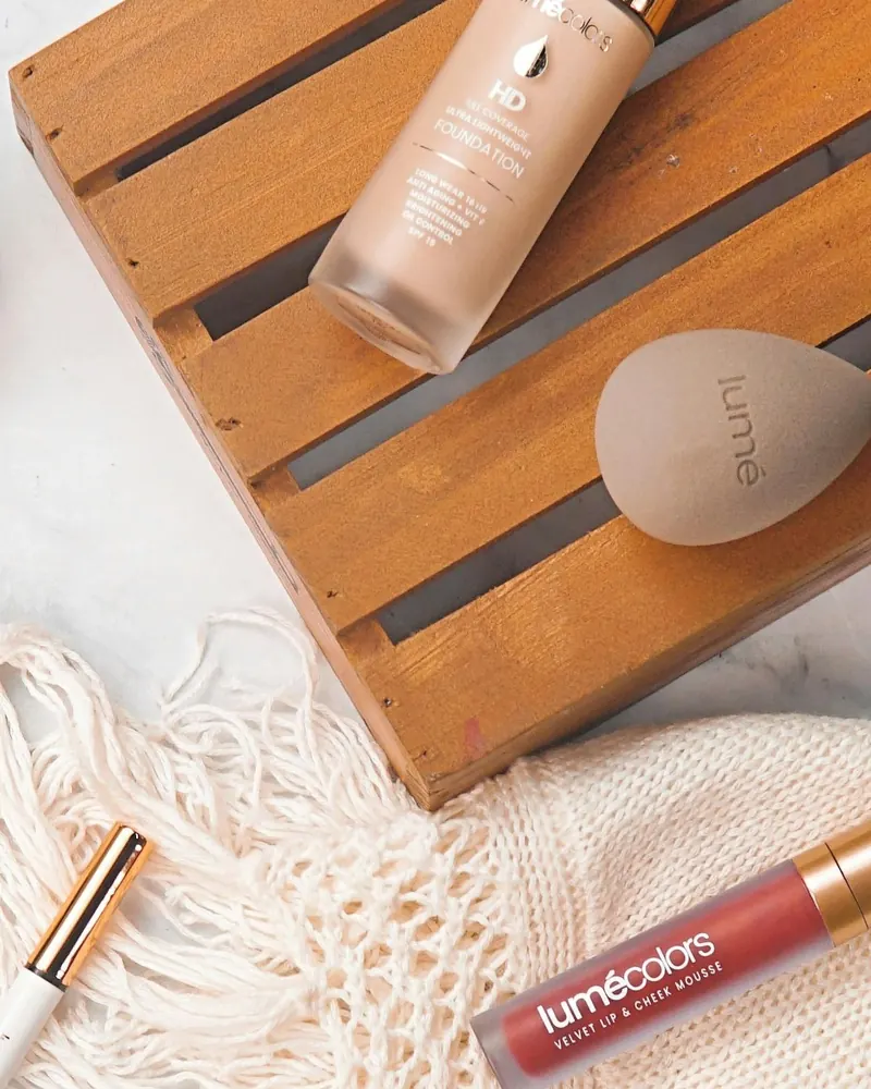 Flat-lay produk Lumécolors: HD Foundation di atas palet kayu, beauty blender, Lip & Cheek Mousse dan Velvet Lipcoat di atas kain rajut